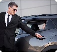 Chauffeur privé de l'équipe Transferts Paris en costume noir et lunettes de soleil ouvrant la porte arrière d'une voiture à une cliente, mettant en avant le service attentionné.